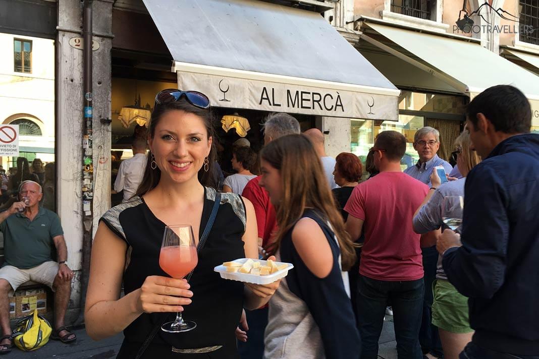 Blogueuse de voyage Biggi Bauer avec une boisson et un en-cas au fromage devant le bar Al Mercà à Venise, avec beaucoup de gens derrière