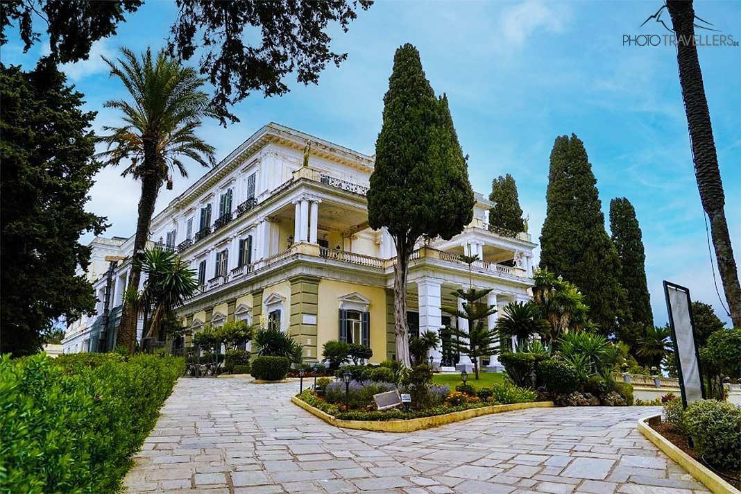 Árboles, palmeras y terrazas de piedra rodean la residencia Achilleion pintada en tonos blanco y amarillo
