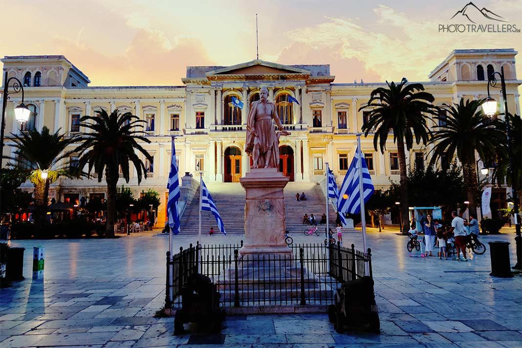Une inscription en pierre entourée de drapeaux grecs devant le bâtiment néoclassique blanc sur la place Miaouli