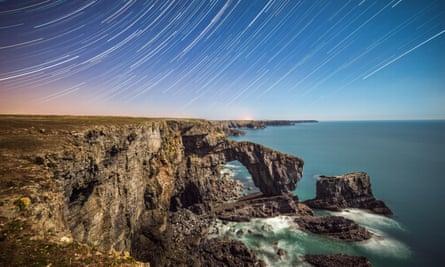 Star trails spiral above the Green Bridge of Wales, illuminated by a near full moon on the south Pembrokeshire coastline