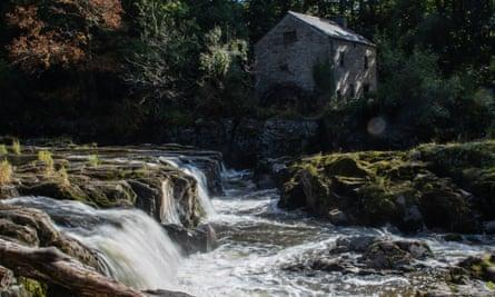 An old stone building in dappled sun beside a stairway of river rapids 
